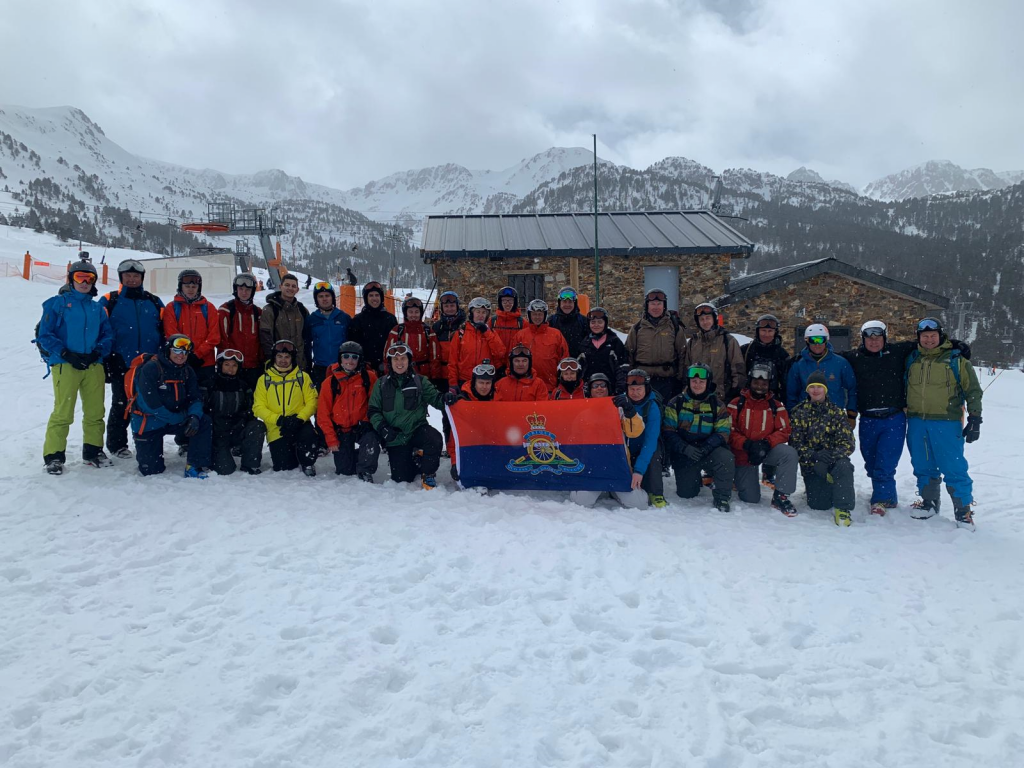 A group of reservists, on a mountain ski resort all dressed in ski gear. They are holding up the British Army flag