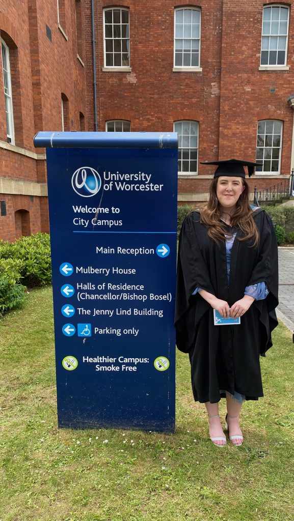 Amy is stood smiling next to the University of Worcester sign. She is dressed in her graduation cap and gown ready to graduate!