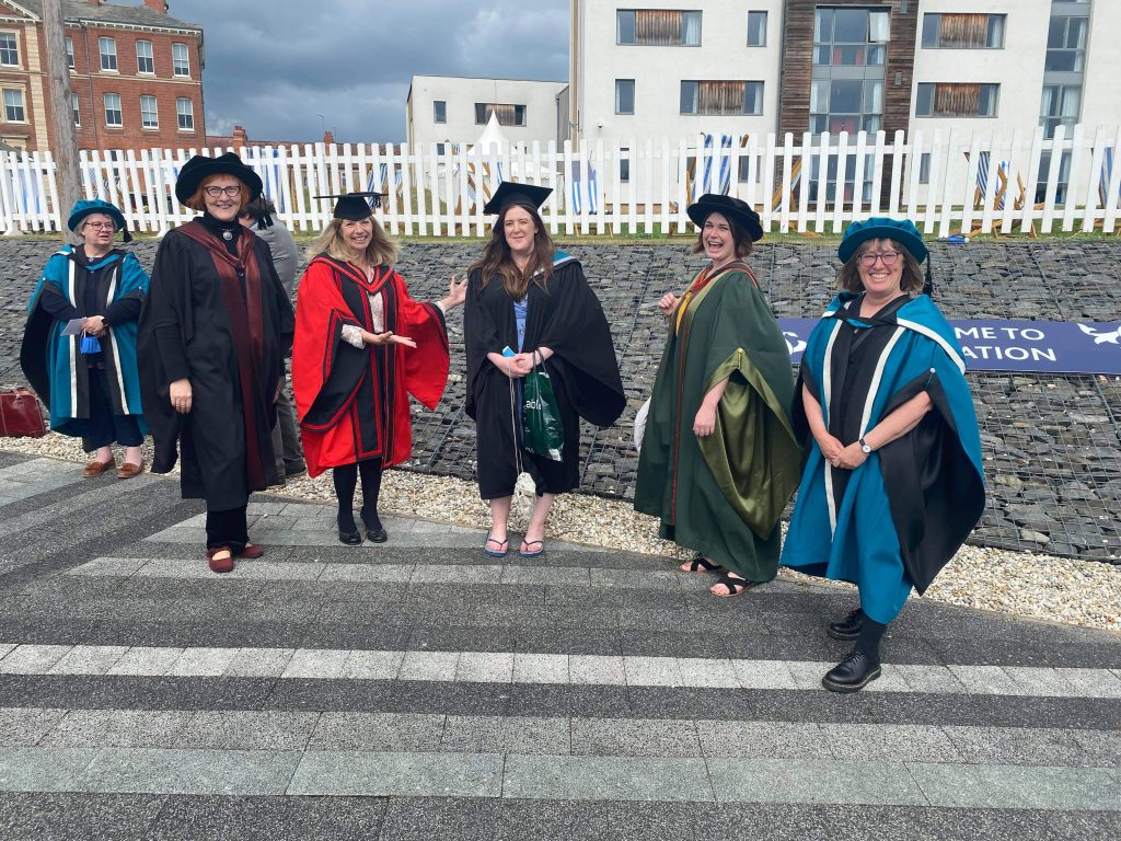 Amy is stood in the middle and is surrounded by her academic lecturers. All are dressed in their graduation cap and gowns however Amy has changed her shoes to a pair of flip flops! 
