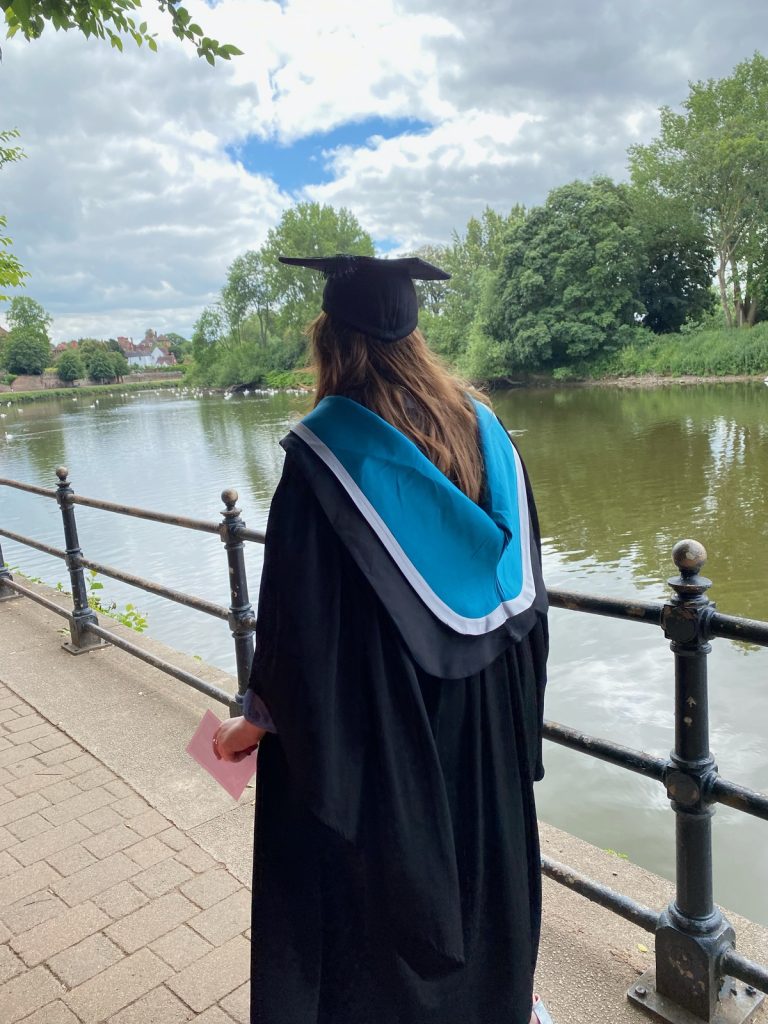 This is an image of Amy walking along the river in her cap and gown. She has her back to us and is looking at the river. 